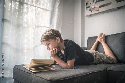 Blond boy barefoot reads book with his face leaning on a hand, student lying down reading the ...