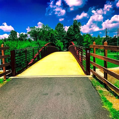 a wooden bridge over a paved road surrounded by green grass and trees on a sunny day