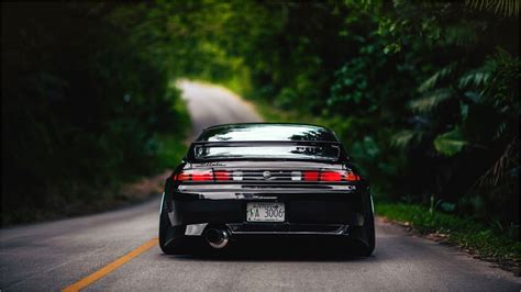 the back end of a black sports car driving down a tree lined road in front of trees