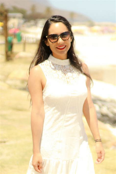 a woman in a white dress and sunglasses walking on the beach with her hand in her pocket