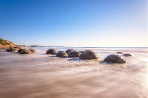 Moeraki Boulders in sea against sky on sunny day at Koekohe Beach, South Island, New Zealand ...