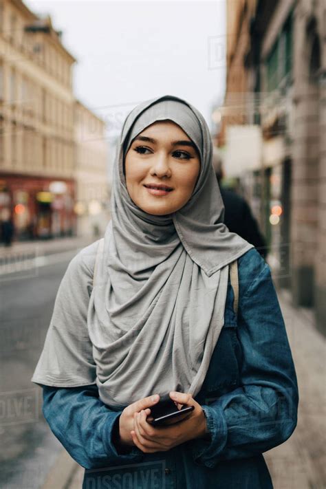 Young Muslim woman wearing hijab walking with mobile phone on sidewalk in city - Stock Photo ...