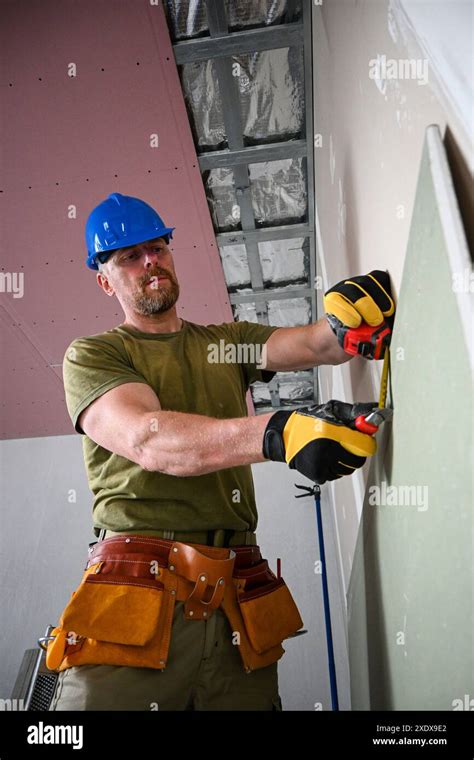 Construction worker wearing tool belt and hard hat measuring plasterboard using tape measure ...