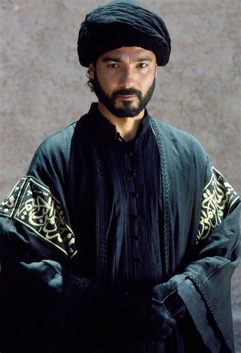 a man with a beard wearing a black and gold outfit standing in front of a stone wall