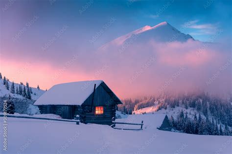 Fantastic winter landscape with wooden house in snowy mountains. Hight mountain peaks in foggy ...