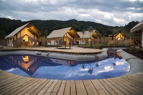 a pool surrounded by wooden decking next to small buildings with lights on them at dusk