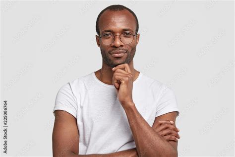 Studio shot of serious black man keeps hand under chin, wears transparet glasses and casual t ...