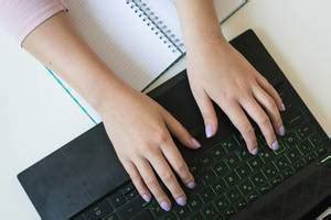 Hands of a woman typing on a black laptop keyboard and a squared notebook with pen seen from ...