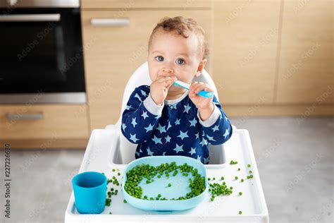 Baby in high chair eating peas with spoon Stock Photo | Adobe Stock