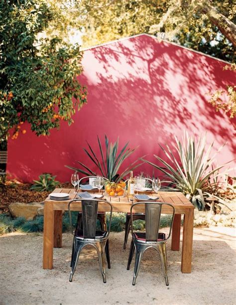 an outdoor dining table with chairs and potted plants in front of a red wall