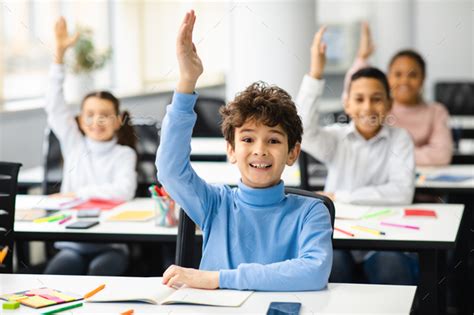 Diverse small schoolkids raising hands at classroom Stock Photo by Prostock-studio