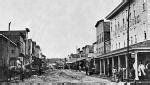 A photograph of a dirt road flanked by two and three-story wooden houses and stores. Visible on ...