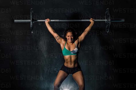 Happy muscular woman win sportswear smiling and lifting heavy barbell over head during intense ...