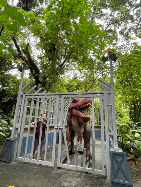 two people standing in front of a gate with a large dinosaur behind it ...