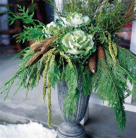 A sleek metal container holds an arrangement of ornamental cabbage, cedar, pine cones, yellow ...