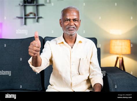 Indian senior old man showing thumbs up sign or hand gesture by looking at camera at home ...