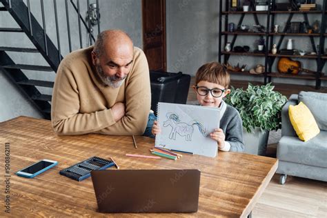 Cute boy showing drawing through laptop sitting by grandfather at home ...