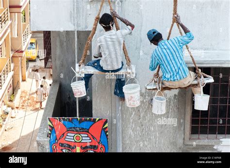 Two indian painters sitting on a wooden seat hanging from ropes painting the side of an ...