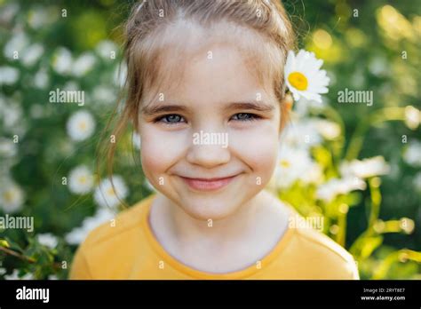 Child portrait of cute little blue-eyed girl with flower of chamomile behind her ear in park or ...