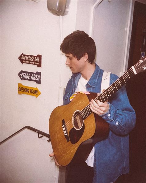 a man holding a guitar in his right hand and wearing a blue jacket on ...