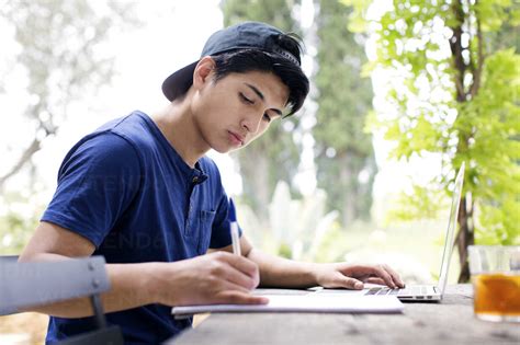 Side view of man writing in book while using laptop at yard stock photo