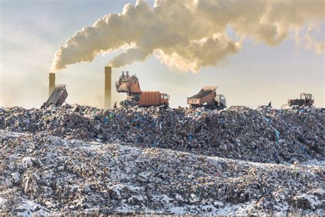 Dump trucks unloading garbage over vast landfill. Smoking industrial stacks on background ...