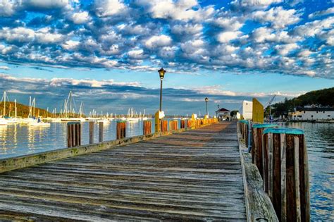 a wooden dock with boats in the water and cloudy skies above it on a ...