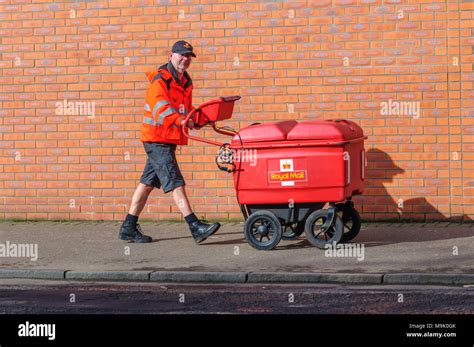 Royal Mail Postman dressed in his uniform wearing shorts, pushing a trolley and delivering the ...