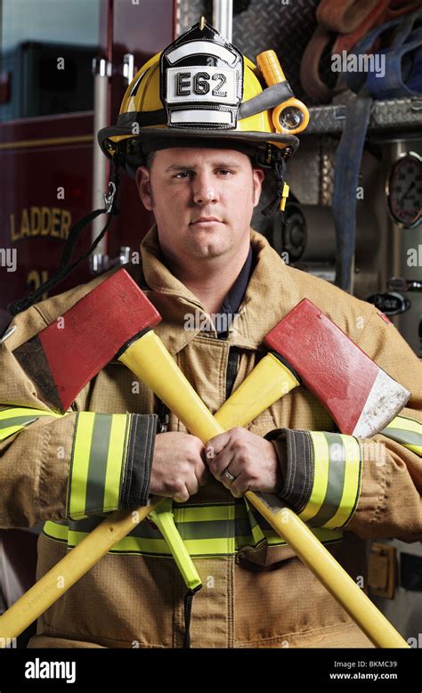 handsome middle age fireman wearing uniform and fire gear holding two axes standing in front of ...