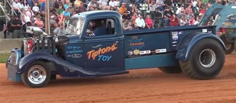 an old blue truck driving down a dirt road in front of a large group of ...