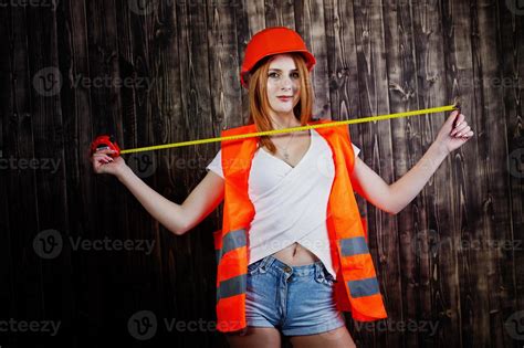 Engineer woman in orange protect helmet and building jacket against wooden background holding ...