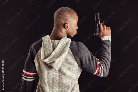 Young handsome black man holding a hand gun, on dark background in studio, seen from the back ...