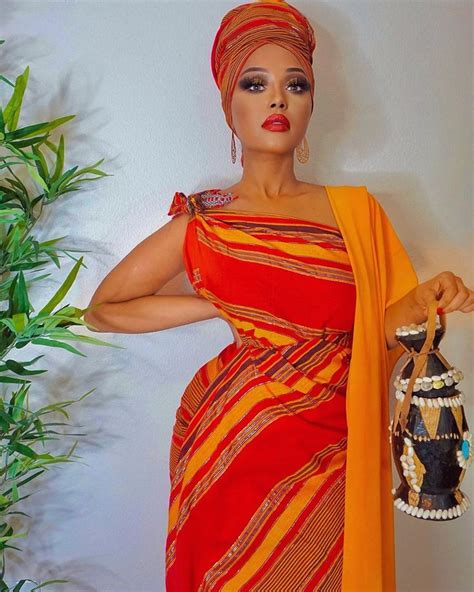 a woman in an orange and red dress holding a purse next to a potted plant