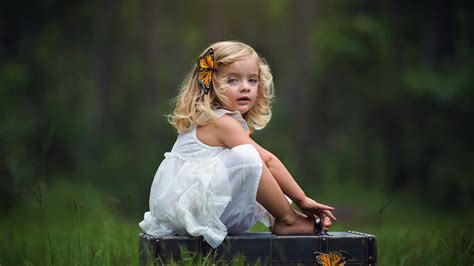 Cute Little Girl Is Sitting On Suitcase Wearing White Dress With Butterfly Clip In Head In Green ...