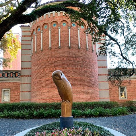 a statue of a bird in front of a brick building with trees and flowers around it