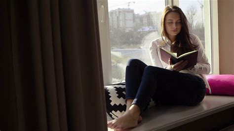 Charming stylish young girl reading a book on the windowsill and looking through the window ...