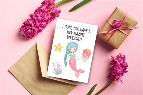 a card with a mermaid on it next to some flowers and a gift box that ...