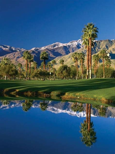 palm trees are reflected in the still water of a golf course with mountains in the background