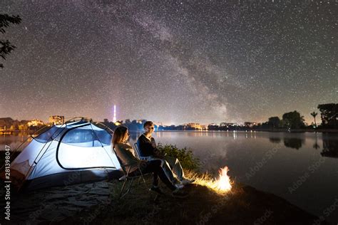 Night camping on lake shore. Couple tourists - man and woman sitting on chairs near campfire and ...