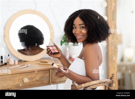 Black woman applying makeup in front of mirror, sitting at dressing table with eye shadow ...