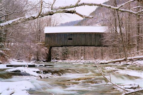 a covered bridge over a small river in the winter with snow on the ground and trees