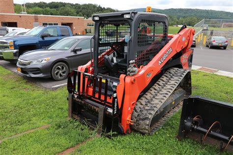 an orange skid steer parked in the grass next to a parking lot with cars