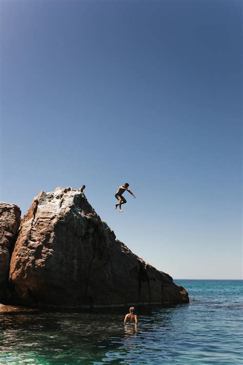 Unrecognizable man jumping off cliff into clear blue water on summer sunny day · Free Stock Photo