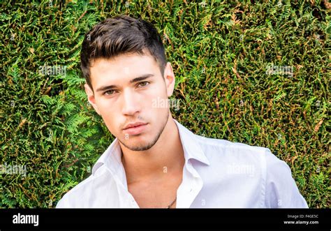 Handsome young man in elegant white shirt standing outdoor in front of green bushy hedge looking ...