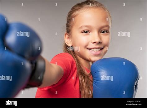 Adorable female child in boxing gloves punching air and smiling while standing against gray ...