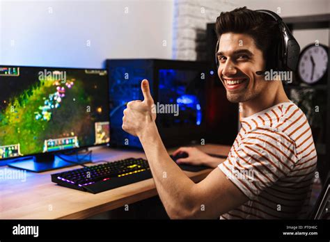 Portrait of joyful gamer guy in headphones playing video games on computer and showing thumb up ...