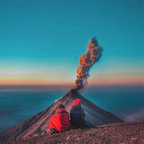 two people sitting on top of a mountain looking at a volcano