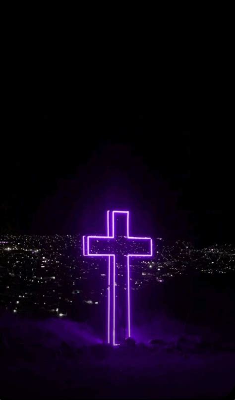 a purple cross is lit up in the night sky with city lights and buildings behind it