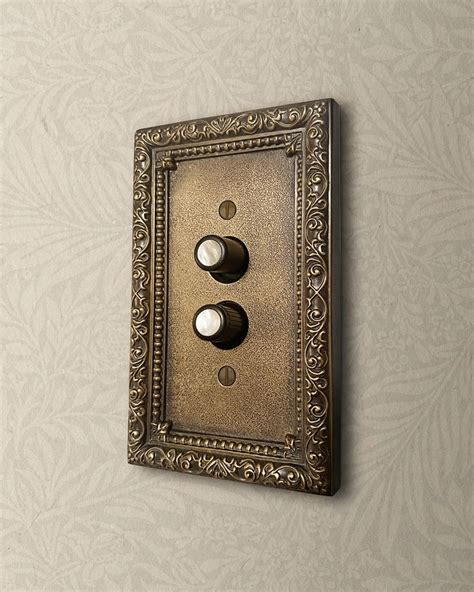 an ornate light switch plate with two dimmers on a wallpapered background