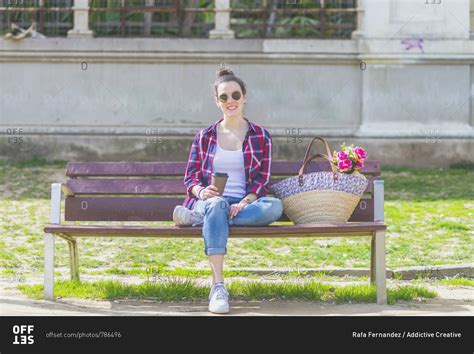Front view of a young hipster woman sitting on a park bench relaxing in a sunny day while ...